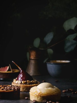 Variety of sweet tartlets with chocolate, caramel, pears, figs with cup of coffee and coffee beans around on black texture table.